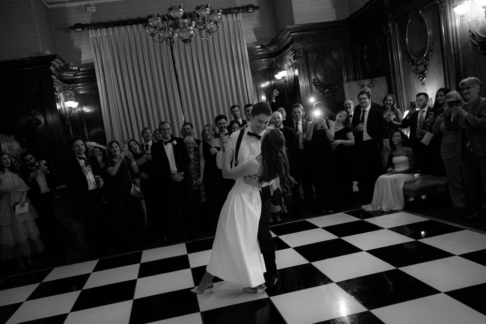 Newlyweds share their first dance on a black and white checkered dance floor surrounded by wedding guests.