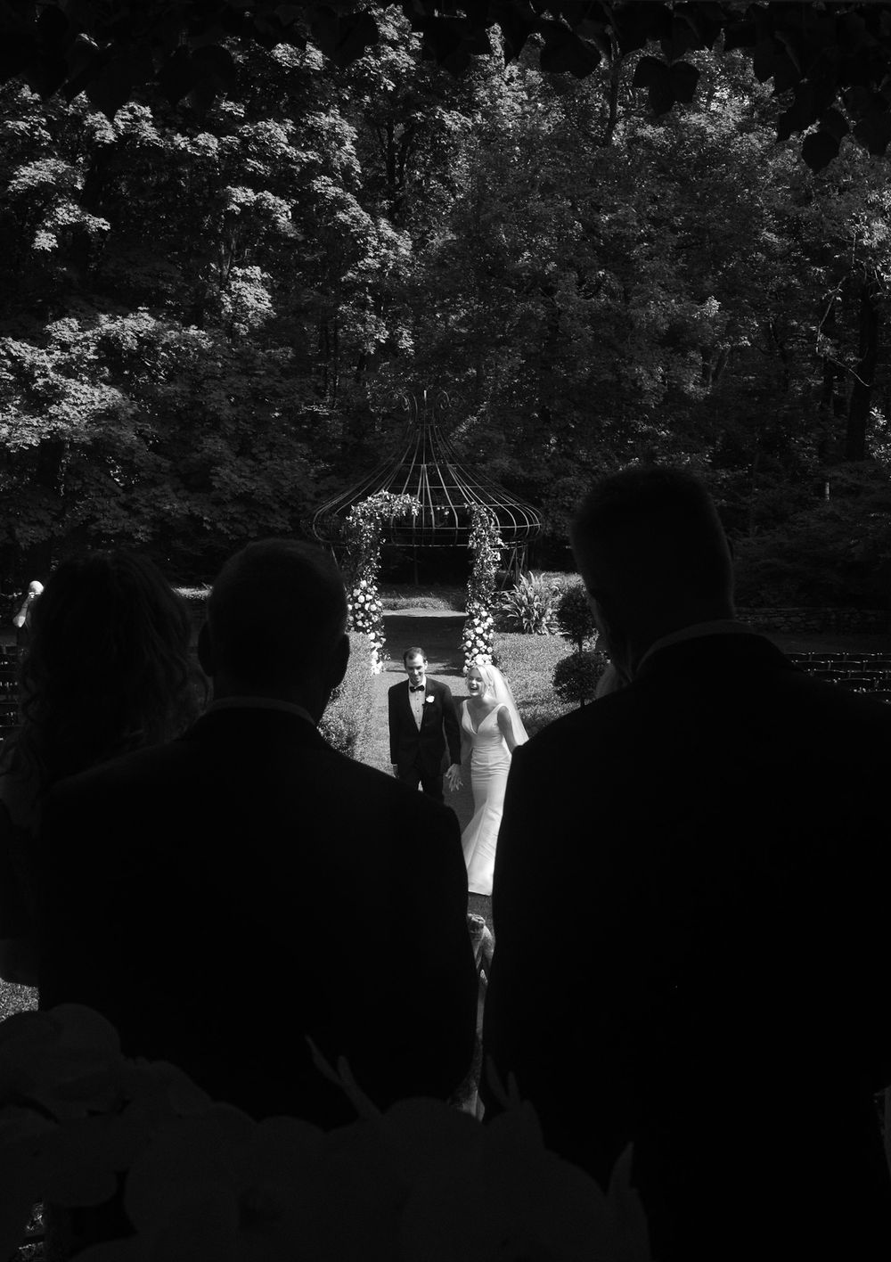 A black and white photograph of an outdoor wedding ceremony under a decorated gazebo surrounded by trees.