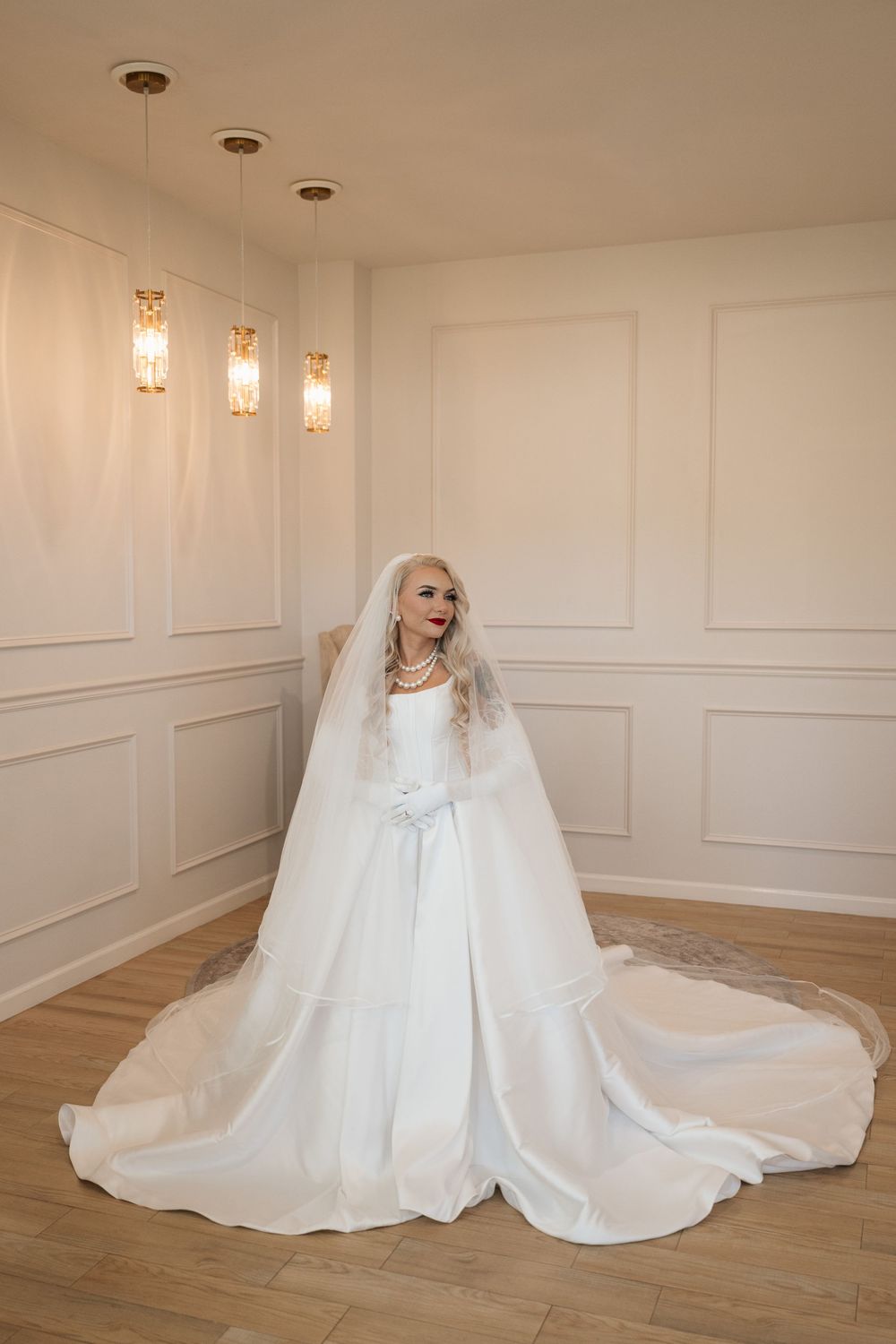Elegant bride in a white ball gown with long veil and gloves posing in a bright white room with pendant lights.