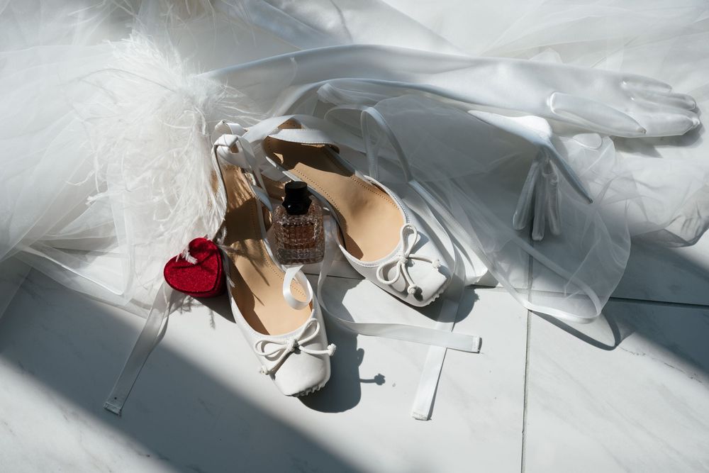 White bridal heels with bow detail, perfume bottle, and red heart charm arranged on white fabric in soft natural light.