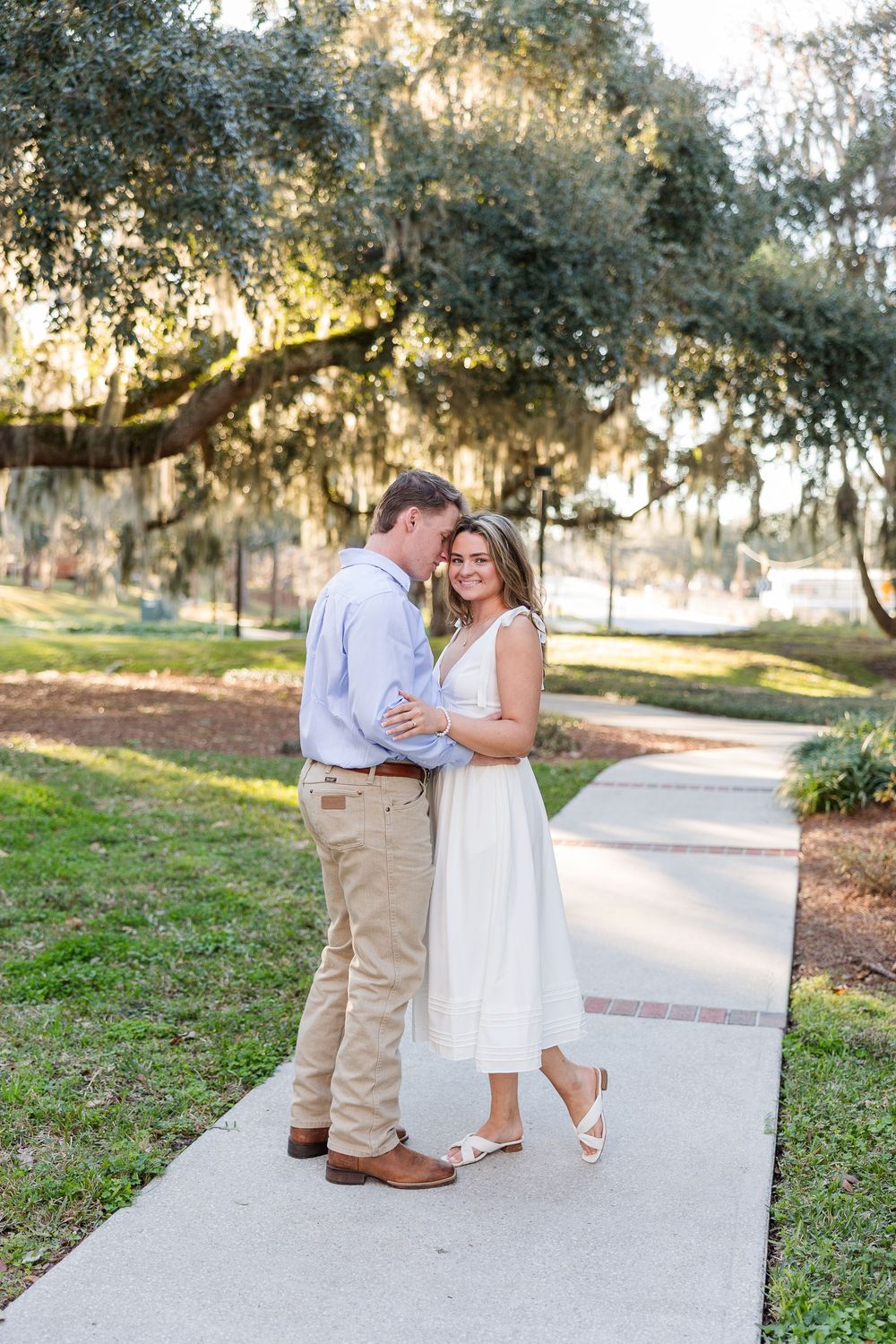 A couple shares a romantic moment on a sunlit path surrounded by Spanish moss draped trees.