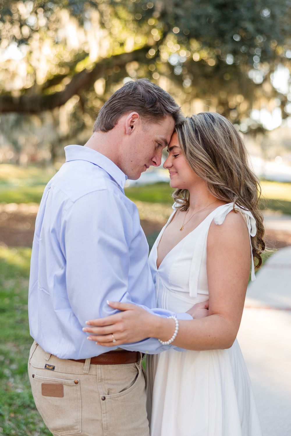 Romantic couple embraces in a sun-dappled outdoor setting wearing casual white and khaki attire.