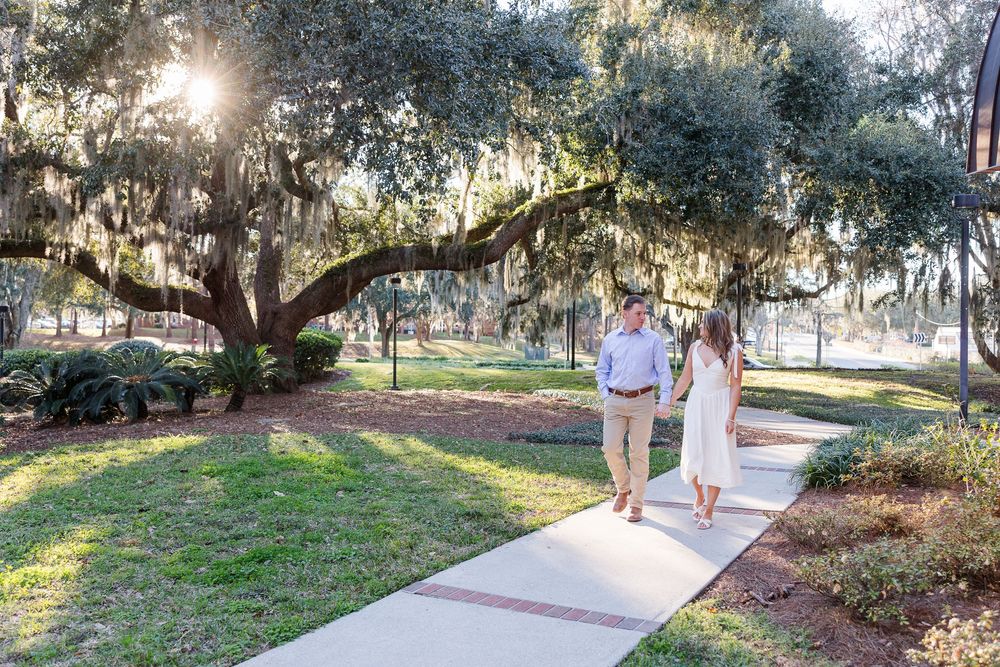 A couple walks hand in hand along a sunlit path beneath Spanish moss-draped oak trees in a romantic park setting.