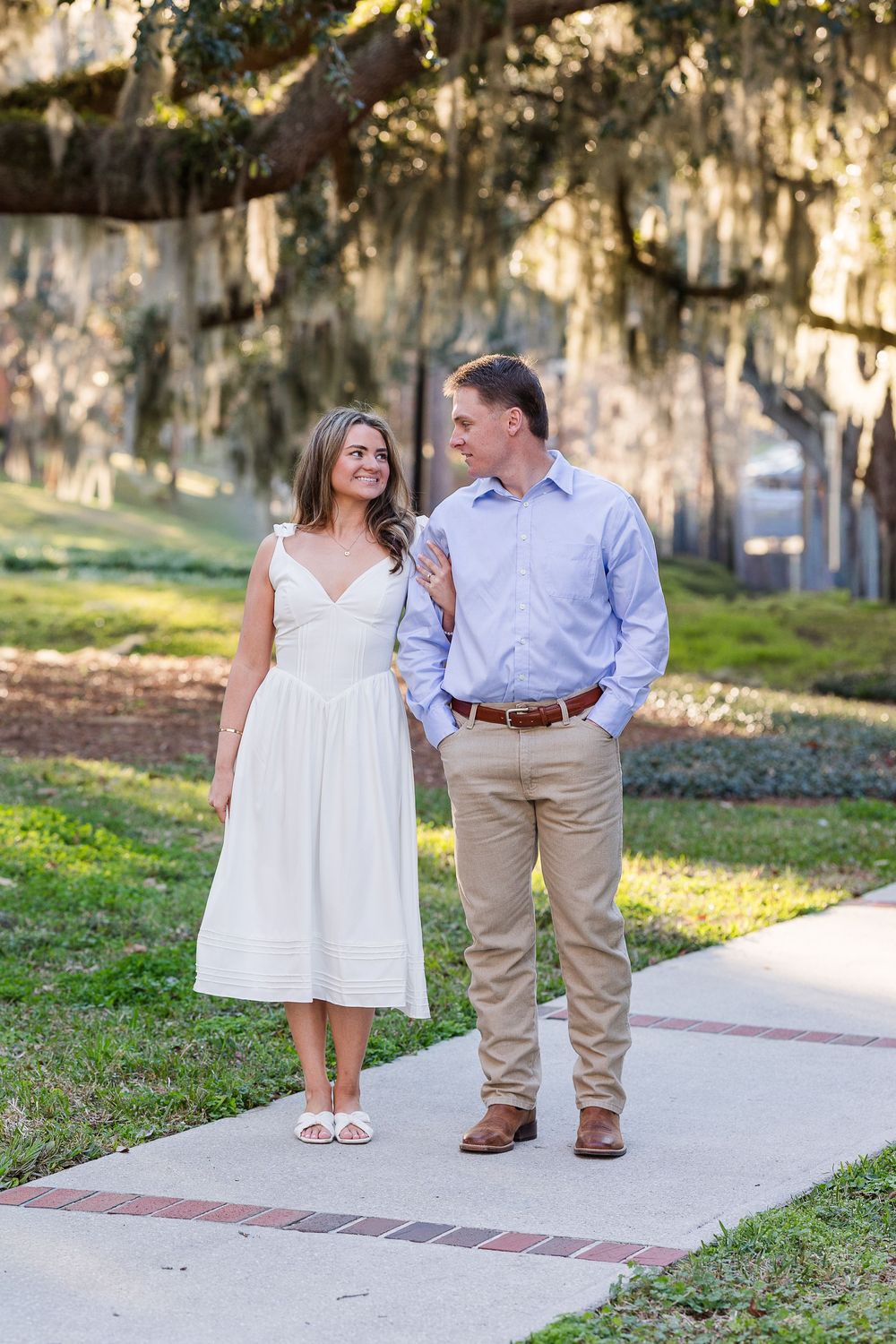 A couple in white and khaki outfits walks together on a pathway surrounded by Spanish moss-draped trees.
