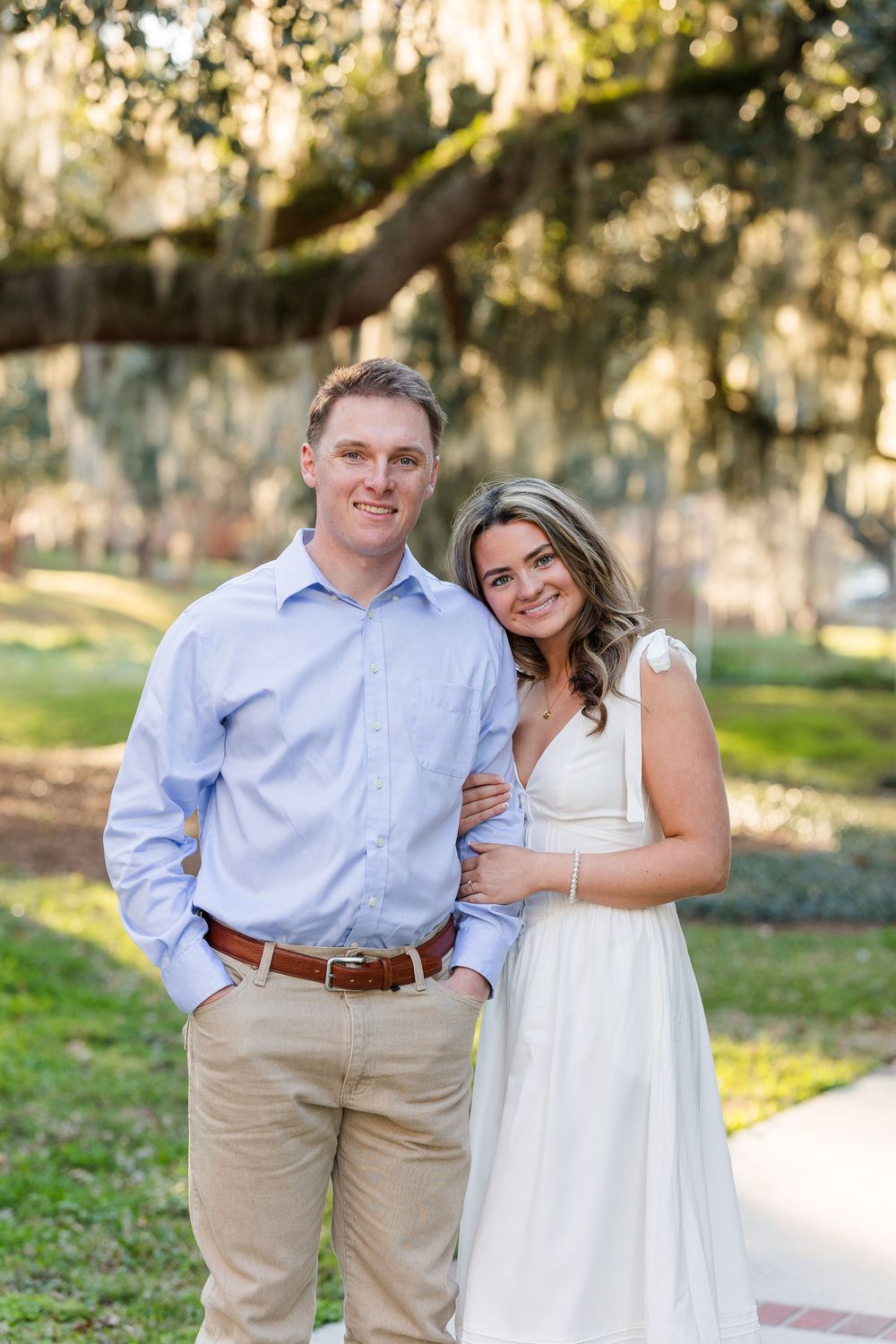 A couple embraces on a walkway in a park with Spanish moss and trees in the background.