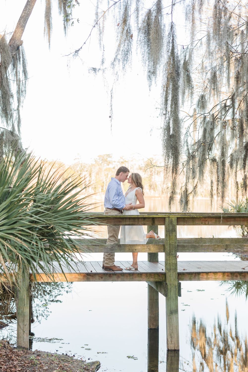 Couple sharing a romantic moment on a wooden dock overlooking a serene lake with Spanish moss hanging from trees.