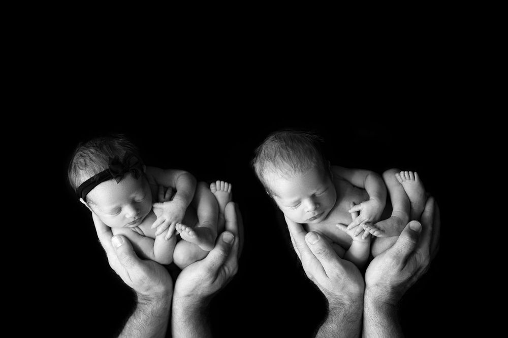 Black and white artistic portrait of two newborn babies cradled in adult hands against dark background.