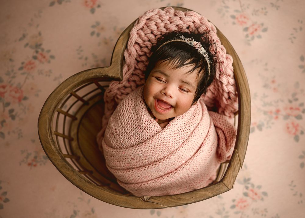 A sleeping baby wrapped in soft pink knit blanket lies peacefully in a heart-shaped wooden basket.