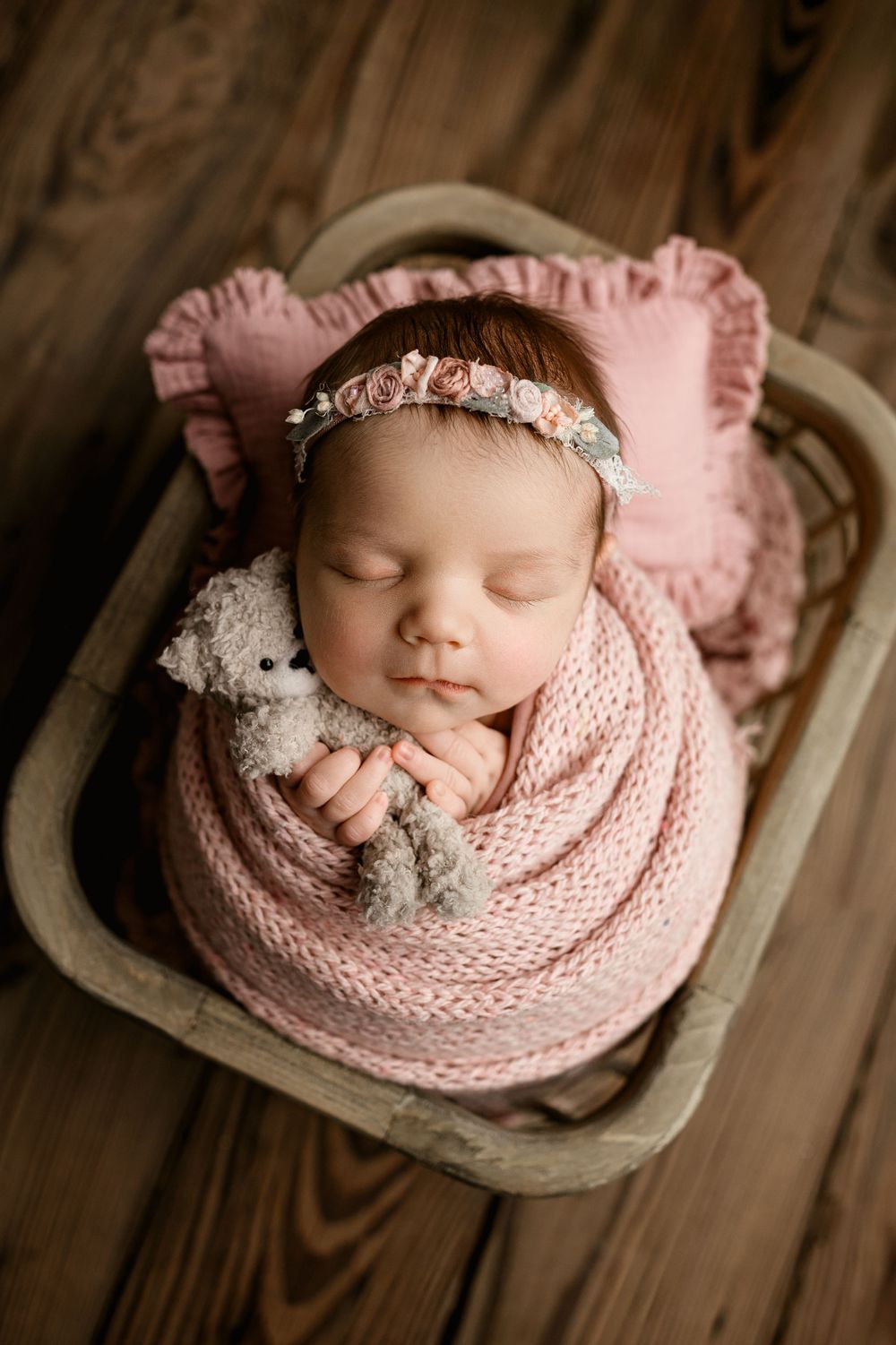 Sleeping newborn baby wrapped in pink knit blanket with delicate headband in rustic wooden setting.