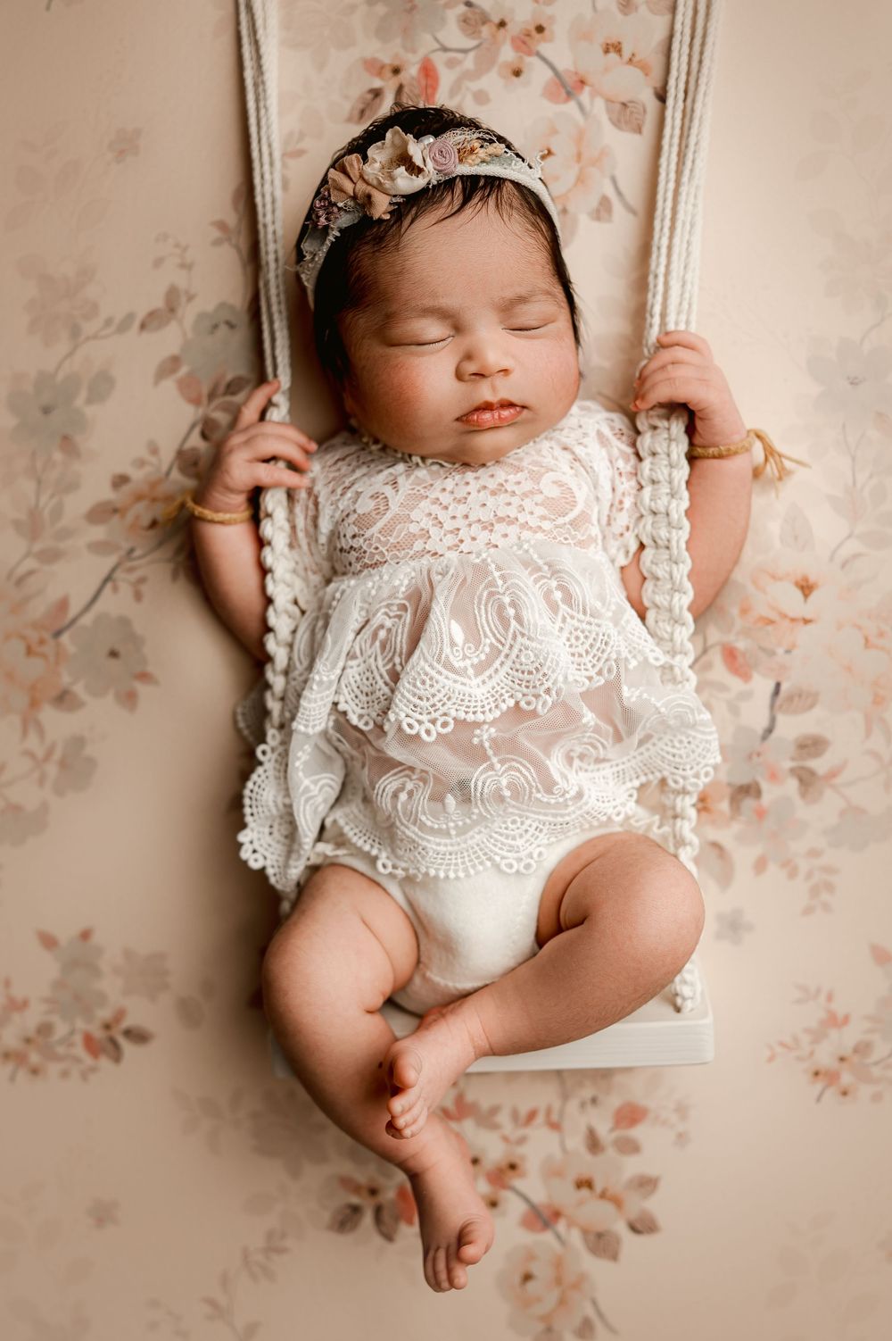 Sleeping newborn baby wearing delicate white lace dress rests peacefully against floral backdrop.