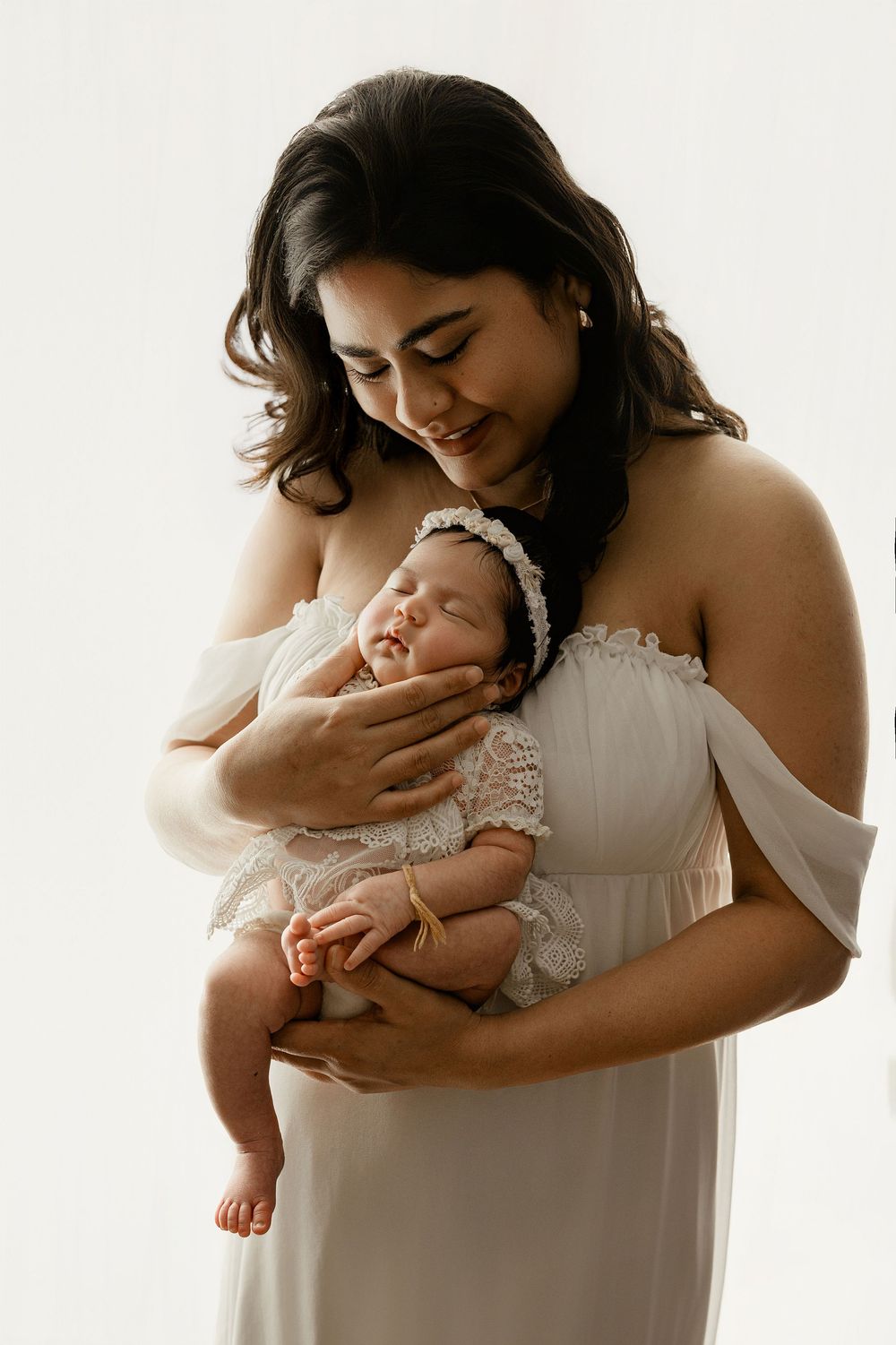 Mother in white dress holds and embraces sleeping newborn baby in soft natural light setting.