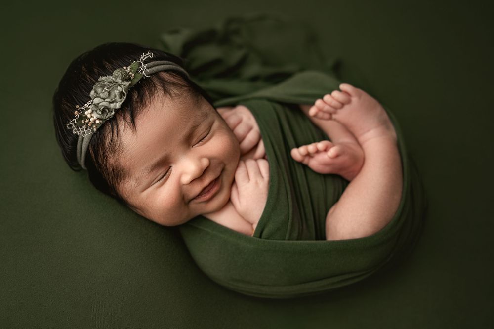Newborn baby wrapped in olive green fabric sleeps peacefully in a dark studio setting.