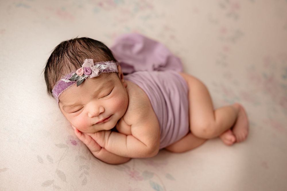 Sleeping newborn baby in purple wrap lying on soft beige blanket with delicate floral headband.