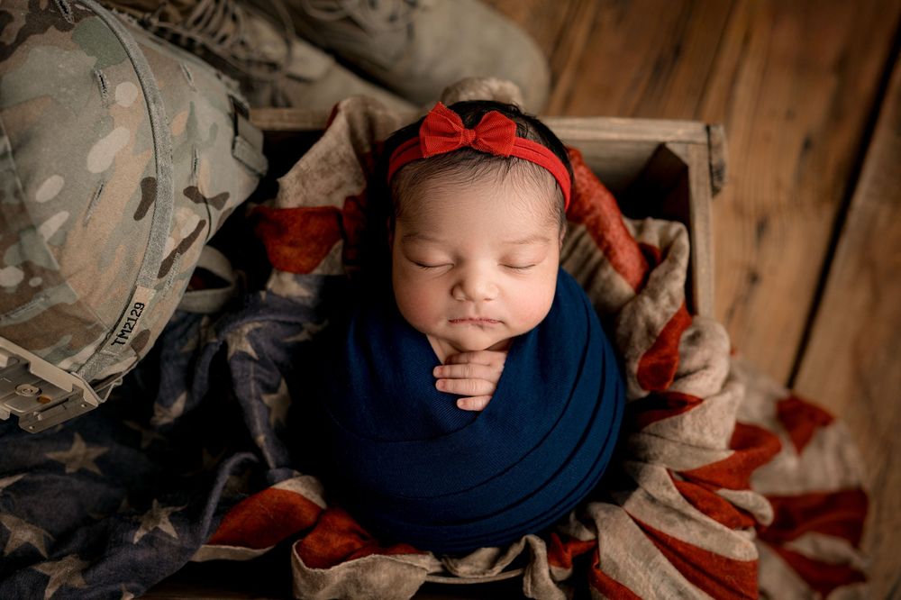 A sleeping newborn wrapped in a navy blue swaddle blanket with American flag decorations in a rustic wooden setting.