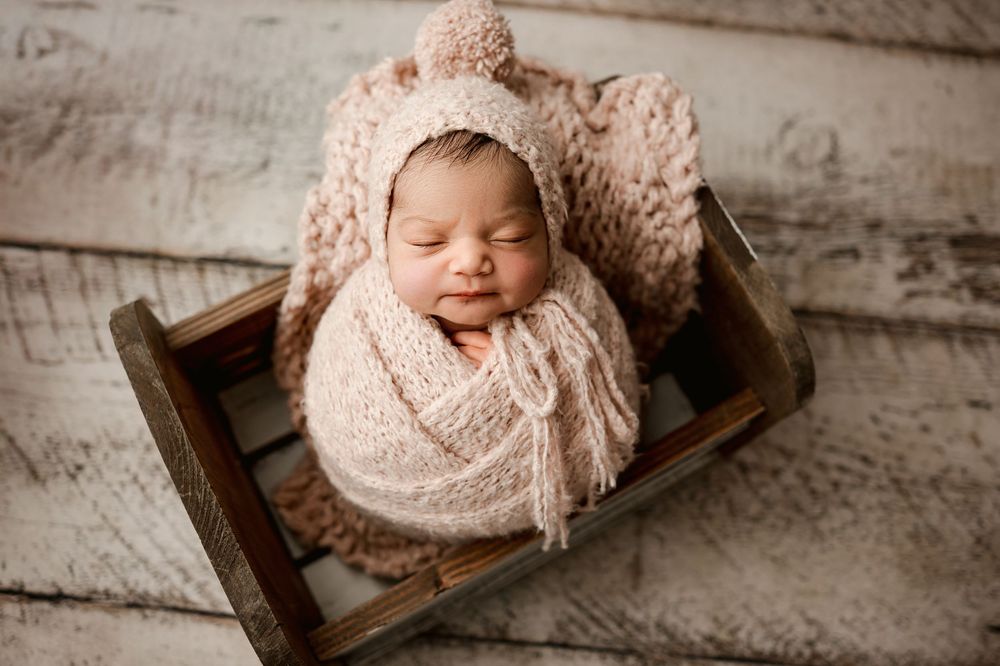 A sleeping newborn swaddled in soft pink knit fabric lies peacefully in a rustic wooden crate.