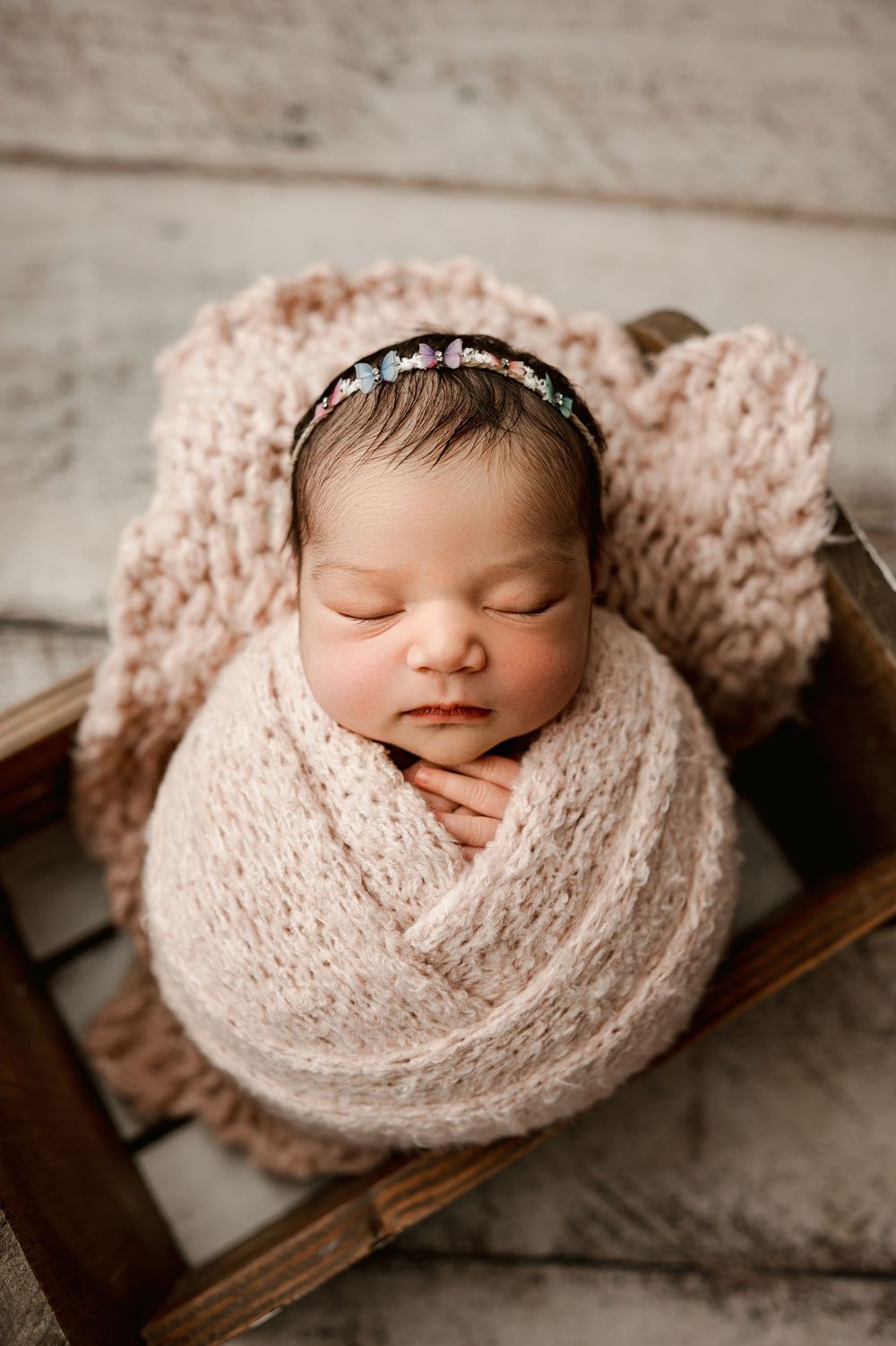Sleeping newborn wrapped in soft pink knit blanket wearing delicate headband rests on wooden surface.