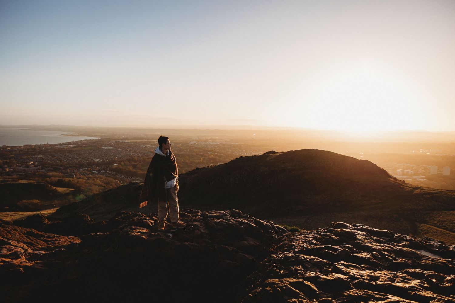 Silhouetted figure stands on rocky mountain peak at sunset overlooking vast desert landscape below. Arthur's Seat