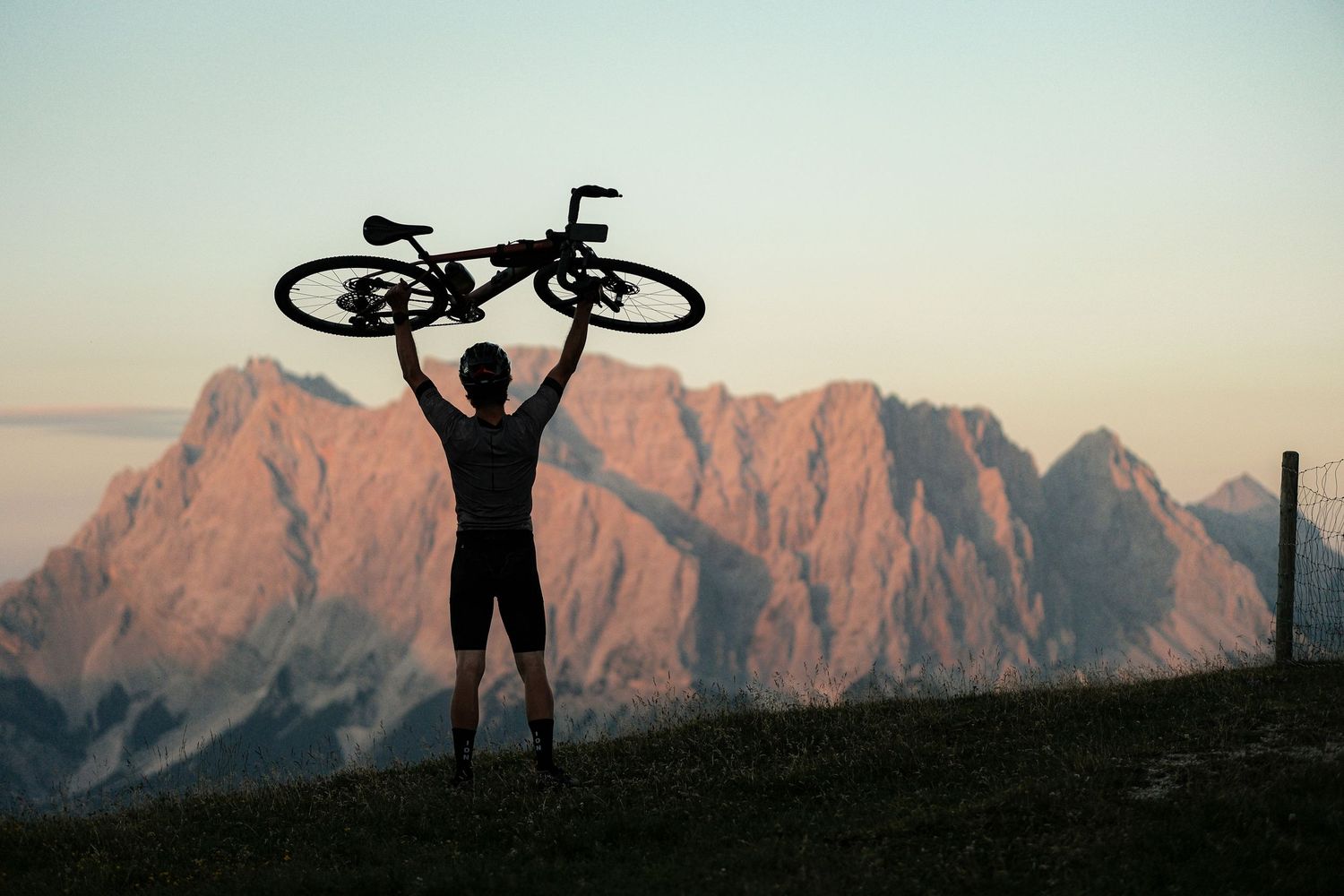 Silhouetted cyclist lifts mountain bike overhead against dramatic mountain peaks at sunset.