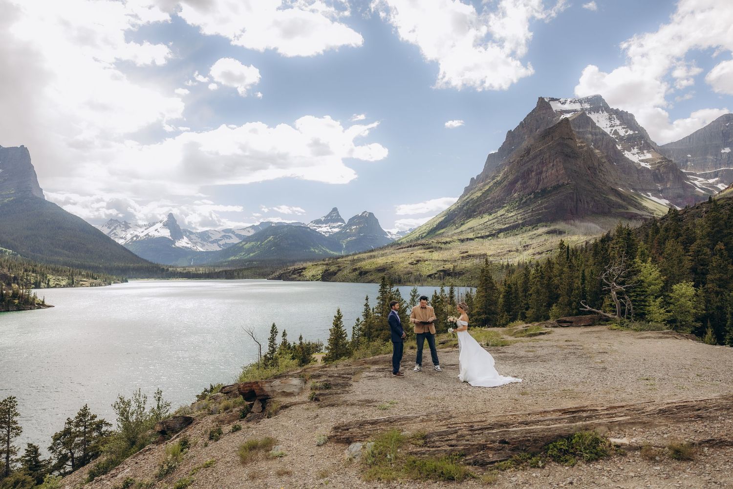 A couple walks through a scenic mountain landscape with tall dry grass and dramatic cloudy skies in the background.