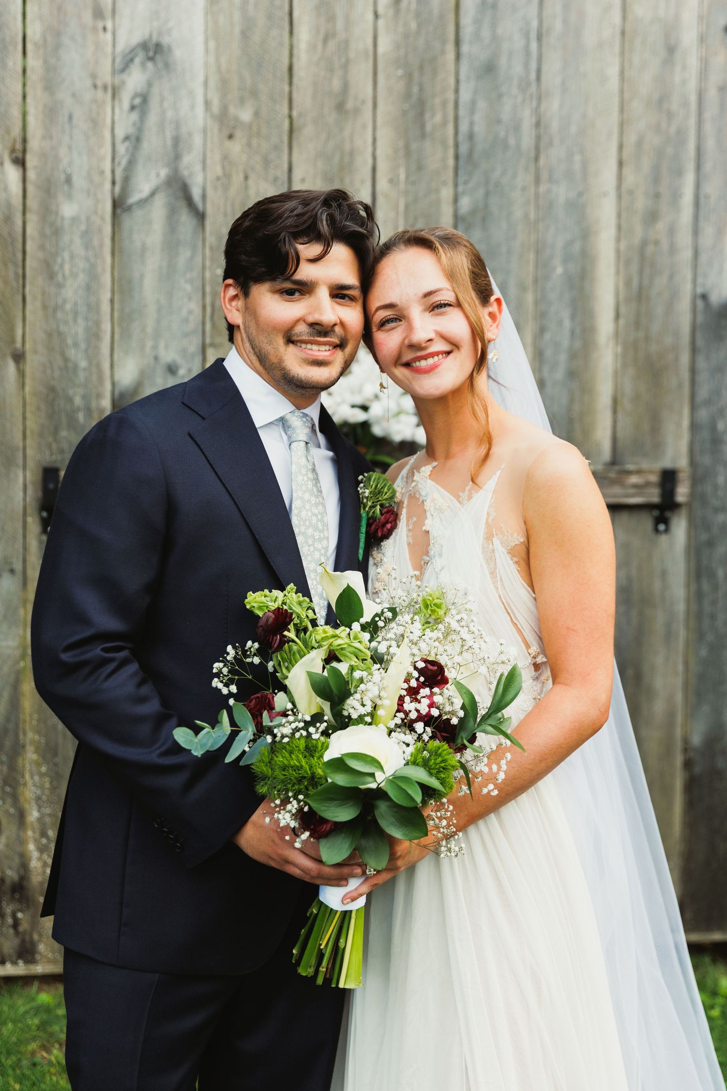 Wedding couple poses with elegant bouquet featuring green foliage against rustic wooden backdrop.