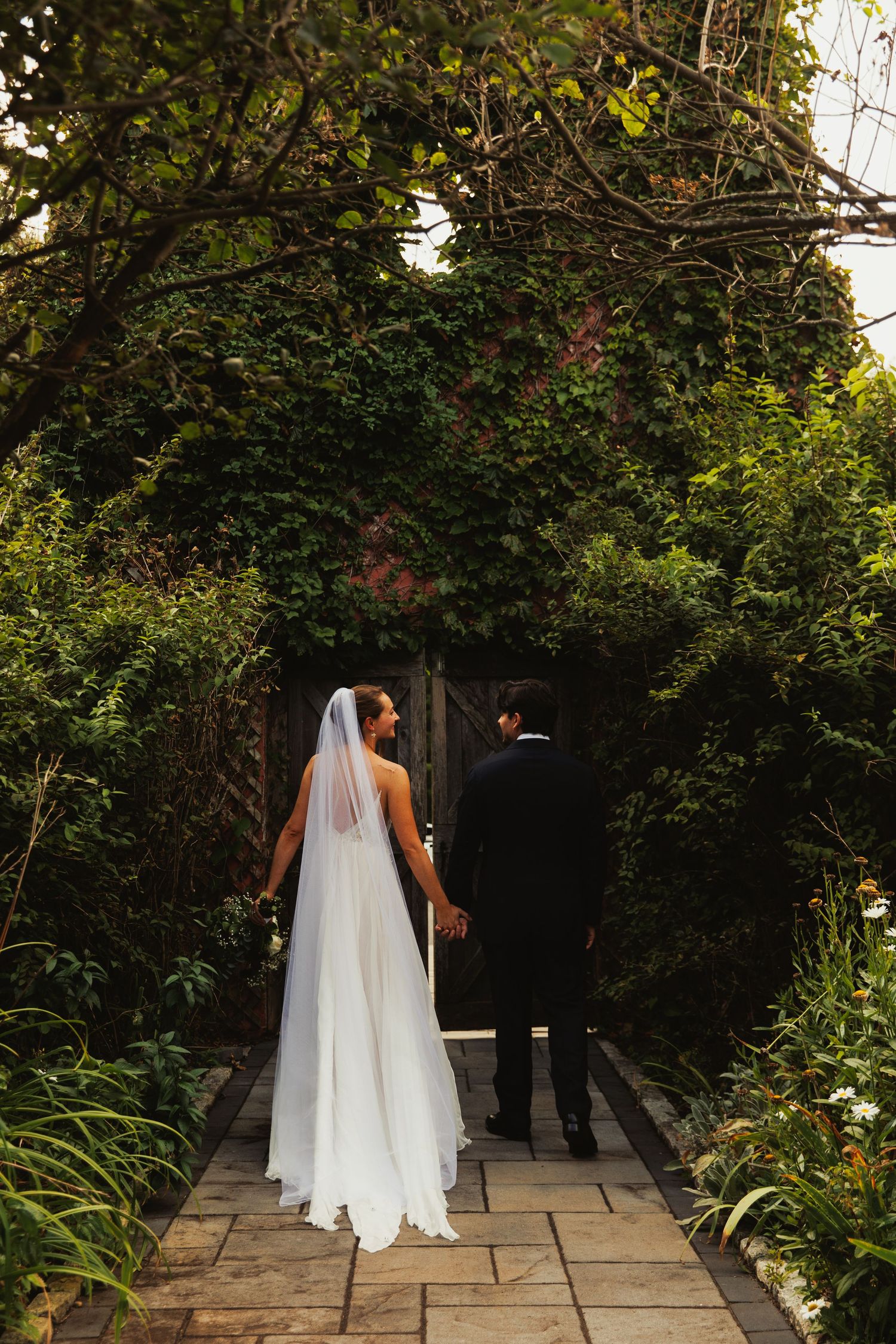 Couple walks along a stone garden path surrounded by lush greenery and flowers.