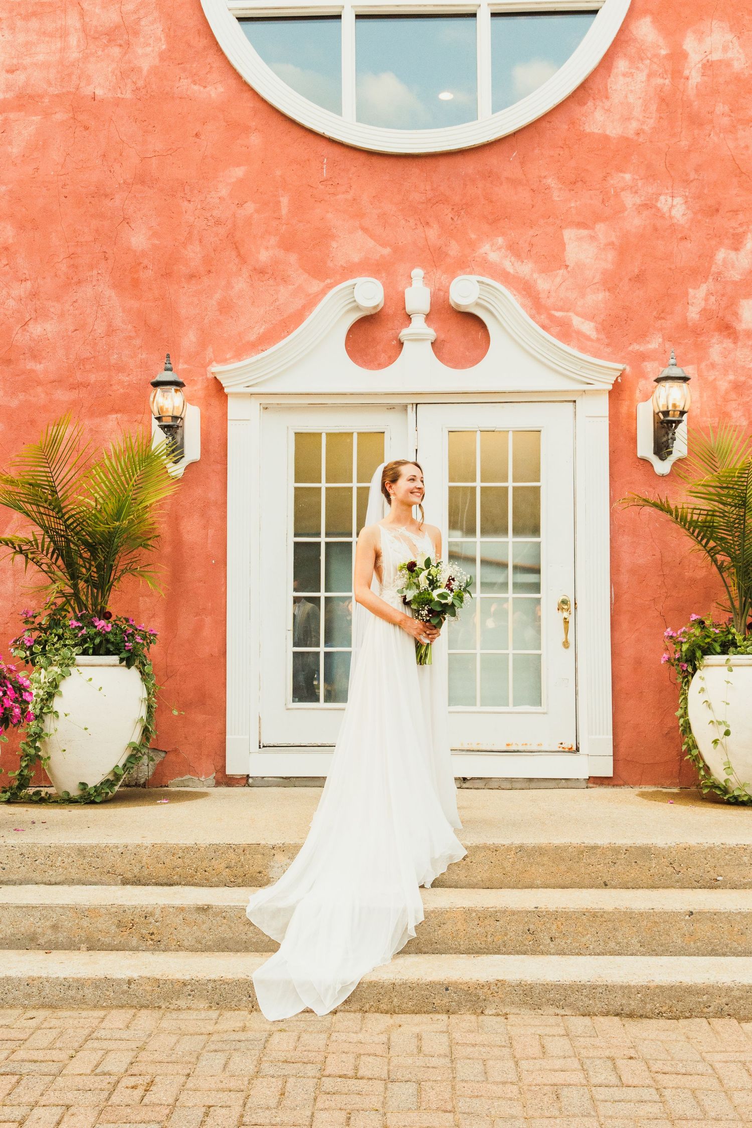 A bride posing against a coral-colored wall with white windows and tropical plants.