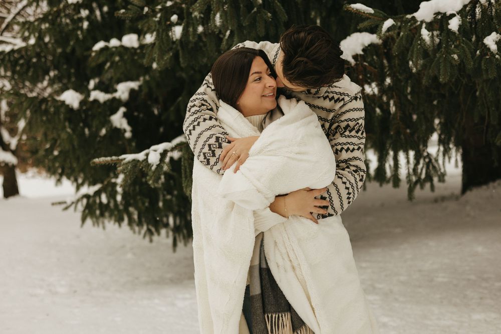 A couple embraces in a snowy winter forest scene while wearing cozy patterned sweaters and white clothing.
