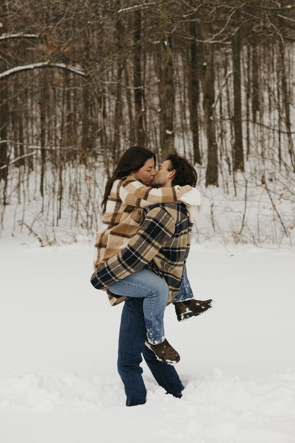 A romantic winter photoshoot shows a couple embracing in a snowy forest wearing plaid and denim.