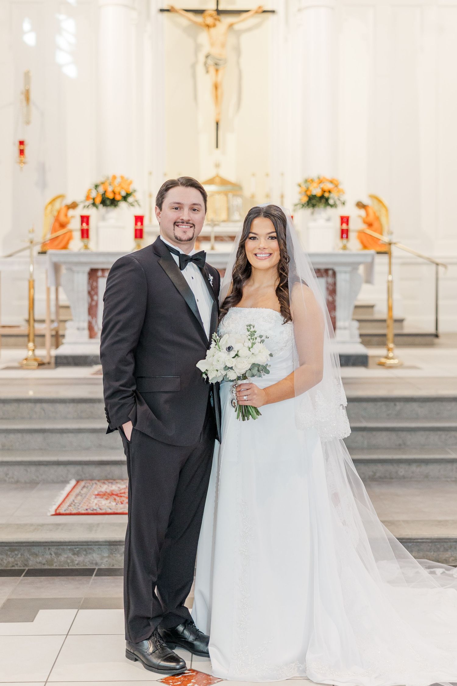 Wedding couple poses together in front of church altar with white flowers and crucifix in elegant ceremony setting.