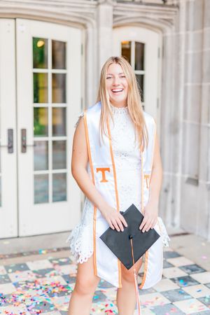 Graduate in white lace dress and orange university stole holds diploma folder on decorative tiled floor.