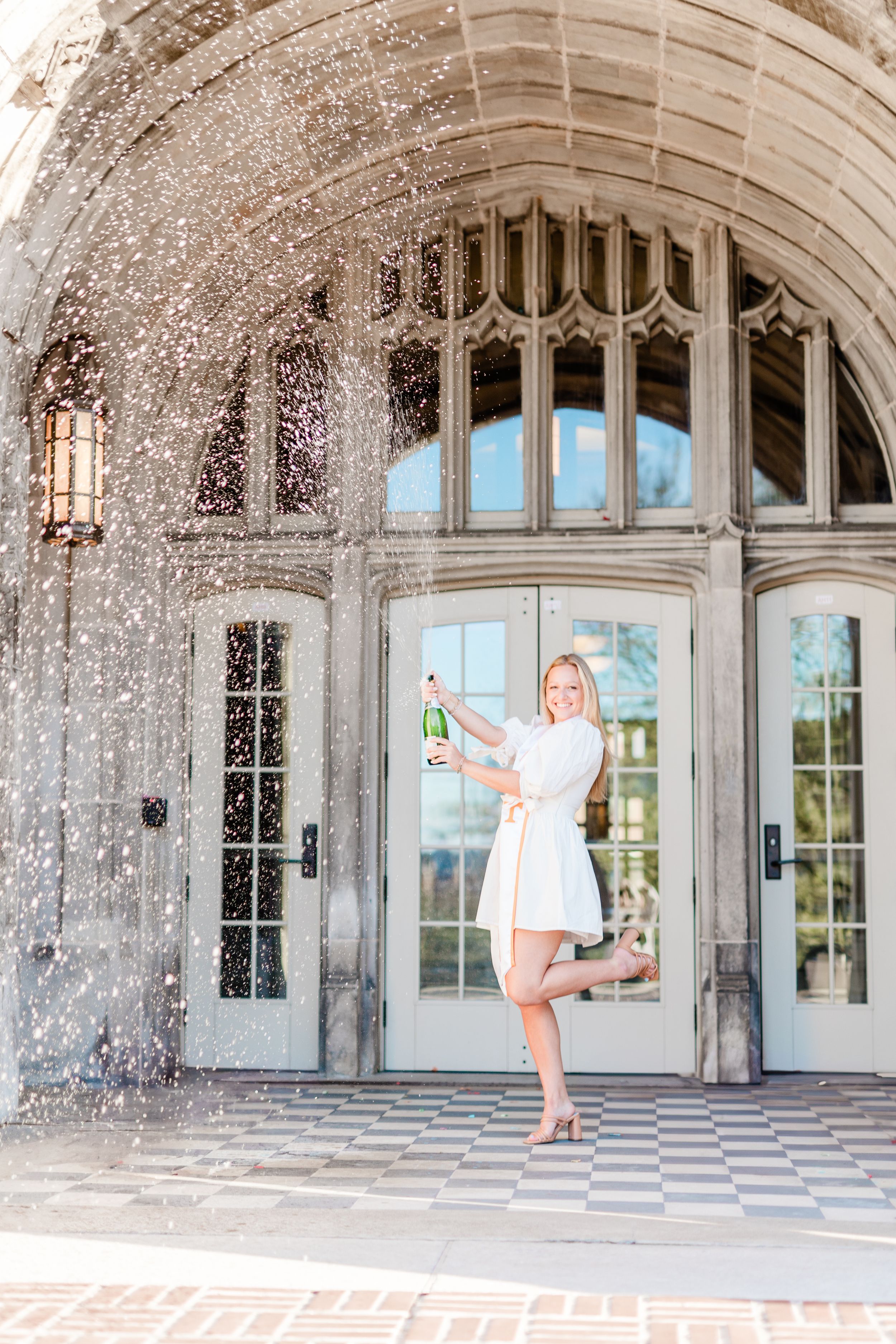 Bride in white dress playfully dances and twirls under an ornate stone archway with sparkling confetti falling around her.
