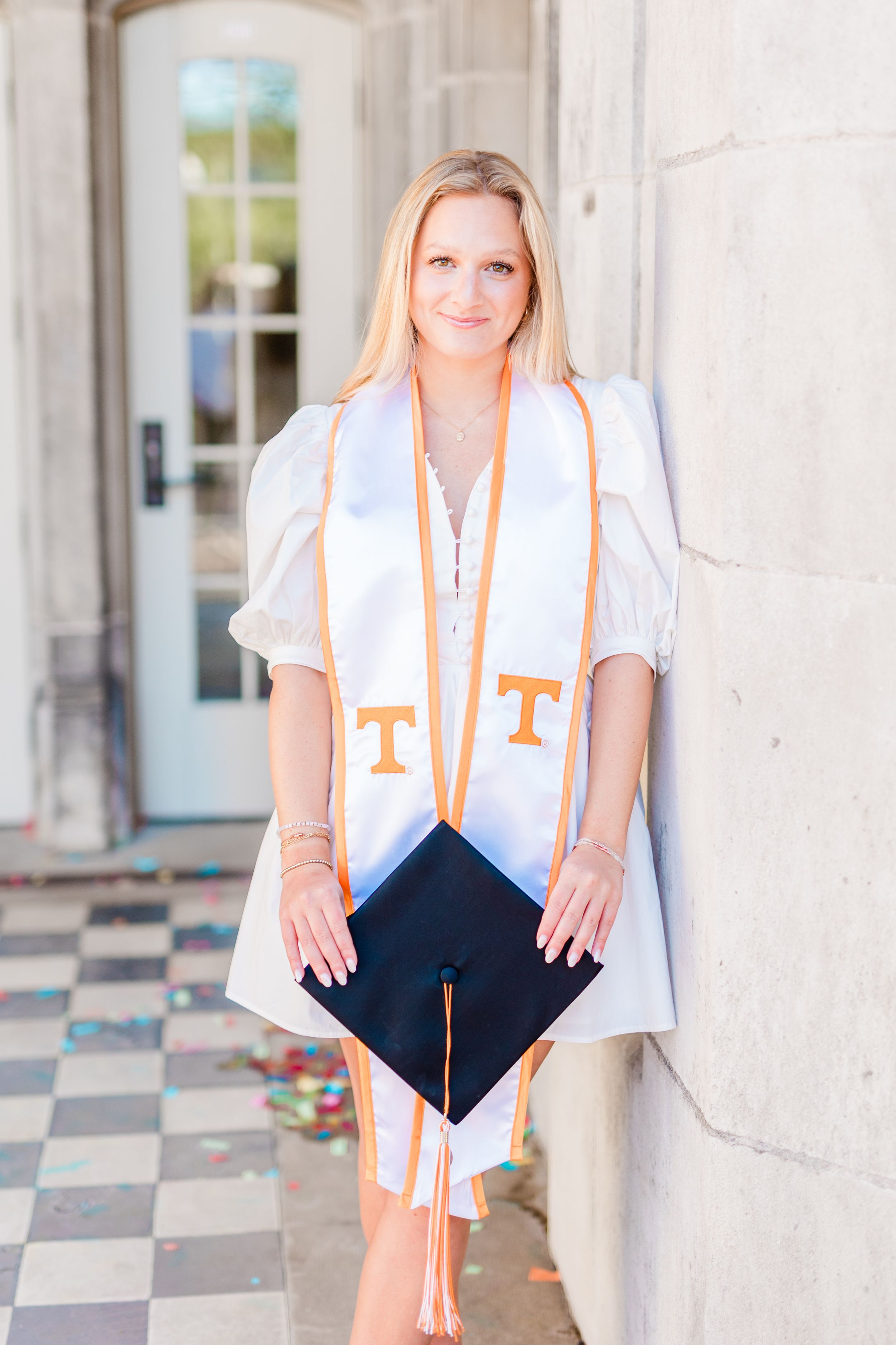 Graduate poses in white shirt and black graduation cap against ornate stone columns in outdoor setting.