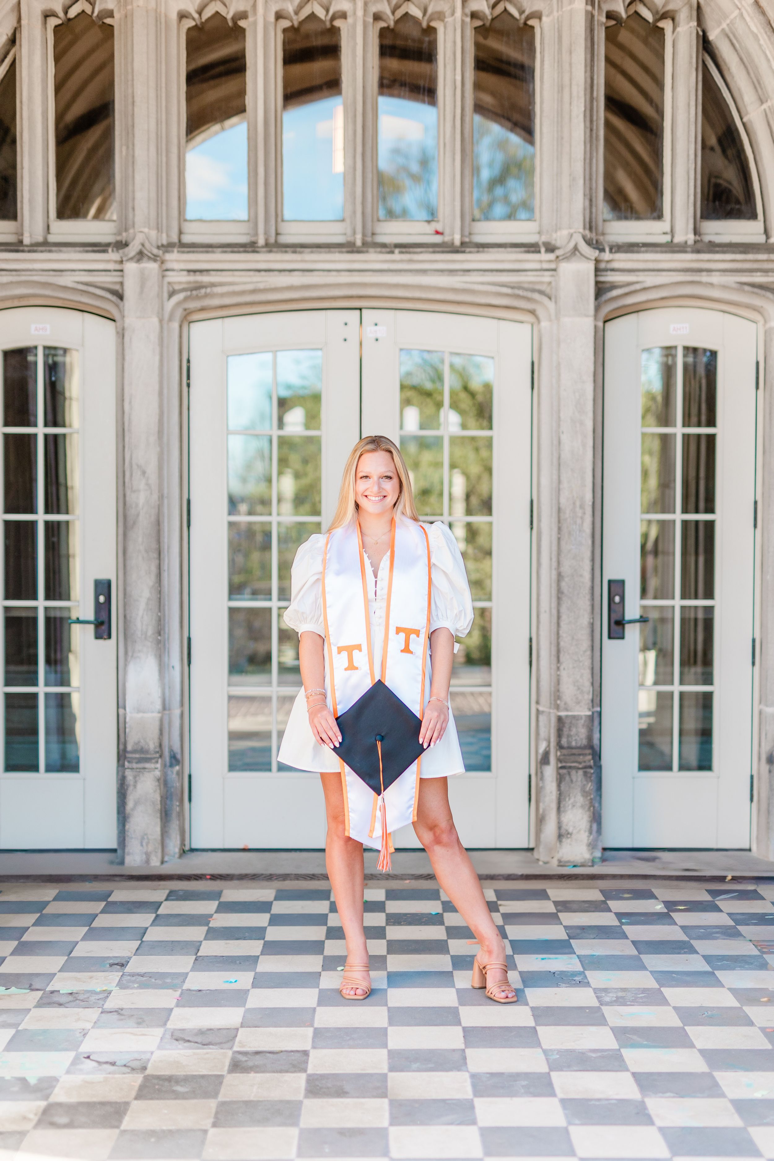 Graduate stands in white dress with graduation cap in front of ornate doorway with checkered floor tiles.