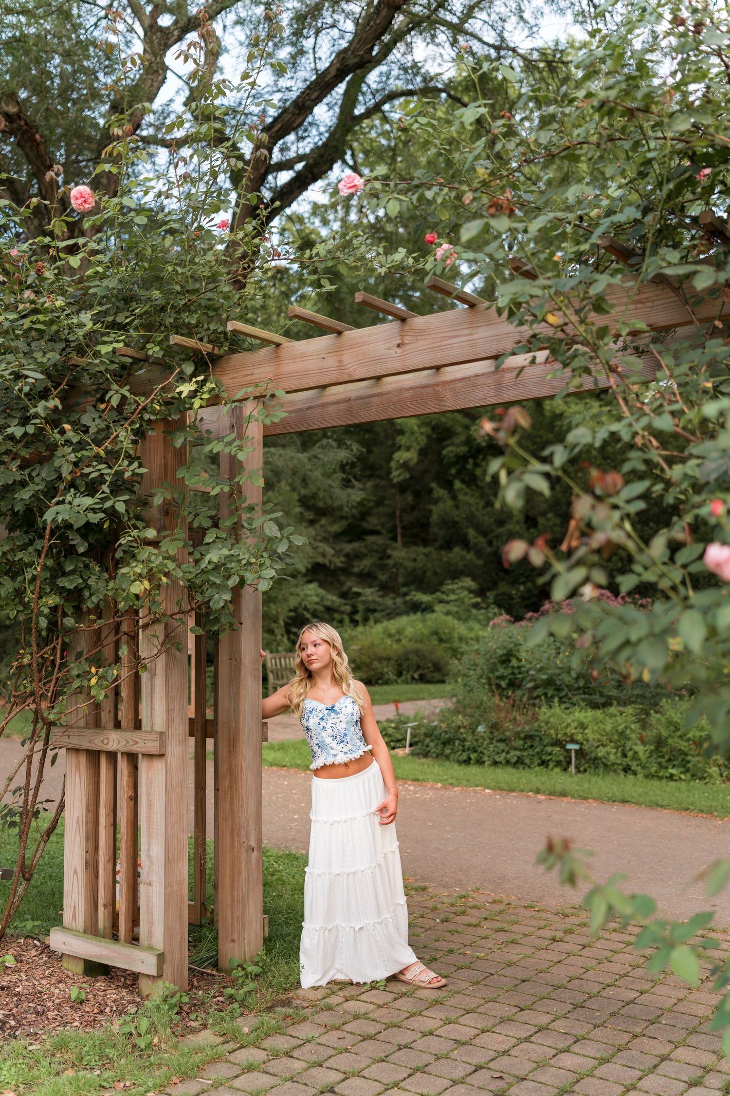 A white dress with embroidered details is photographed in a garden setting with wooden pergola and brick pathway.