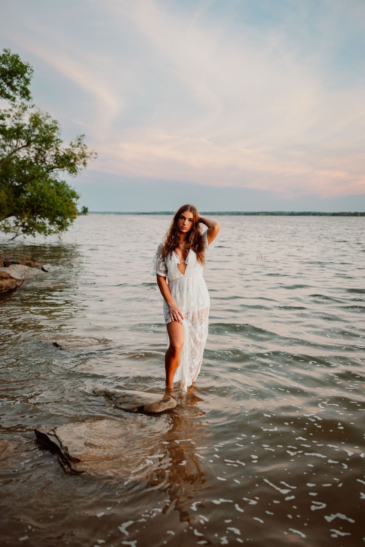A series of photos showing someone in a white dress wading in a serene lake at sunset with trees along the shoreline.