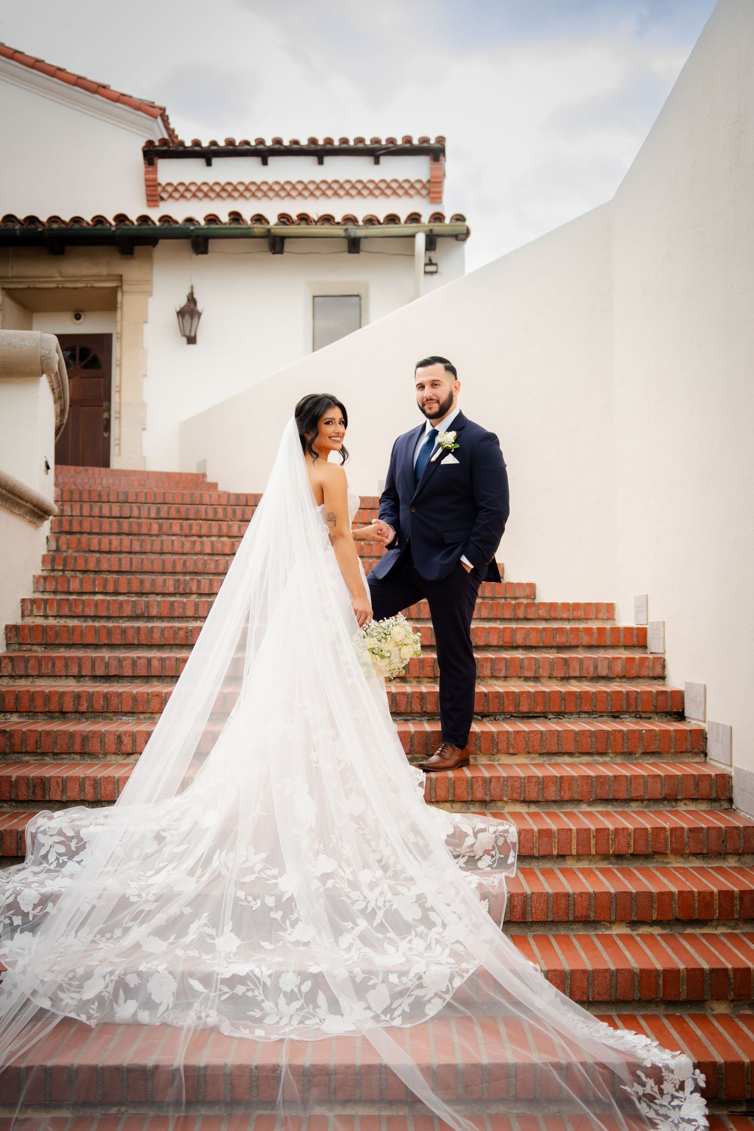 A bride and groom pose on elegant Spanish-style terracotta steps with a flowing white veil creating a romantic scene.