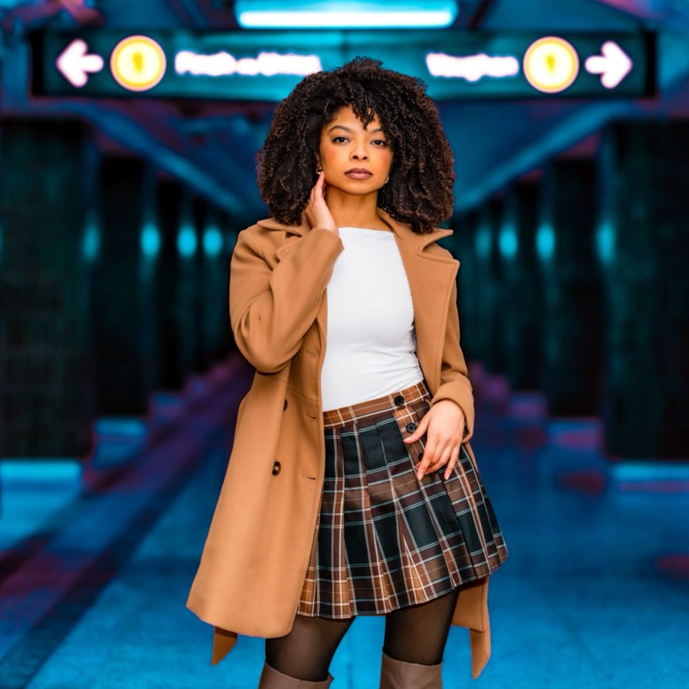 Young Black woman in camel coat and plaid skirt poses in a vibrant blue subway station.