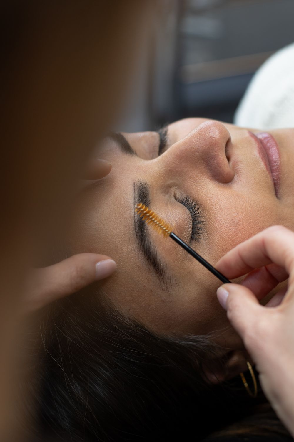 Esthetician applying orange mascara wand to woman's eyelashes during a lash treatment at a beauty salon.