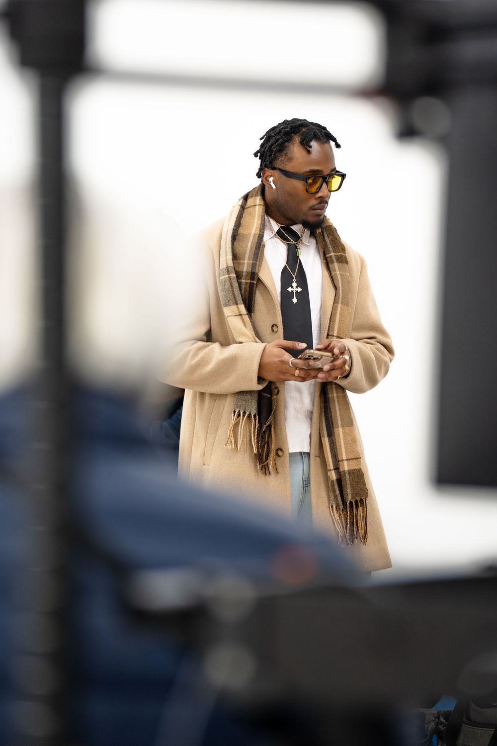 A person in a beige coat and scarf checks their phone while standing outdoors in an urban setting.