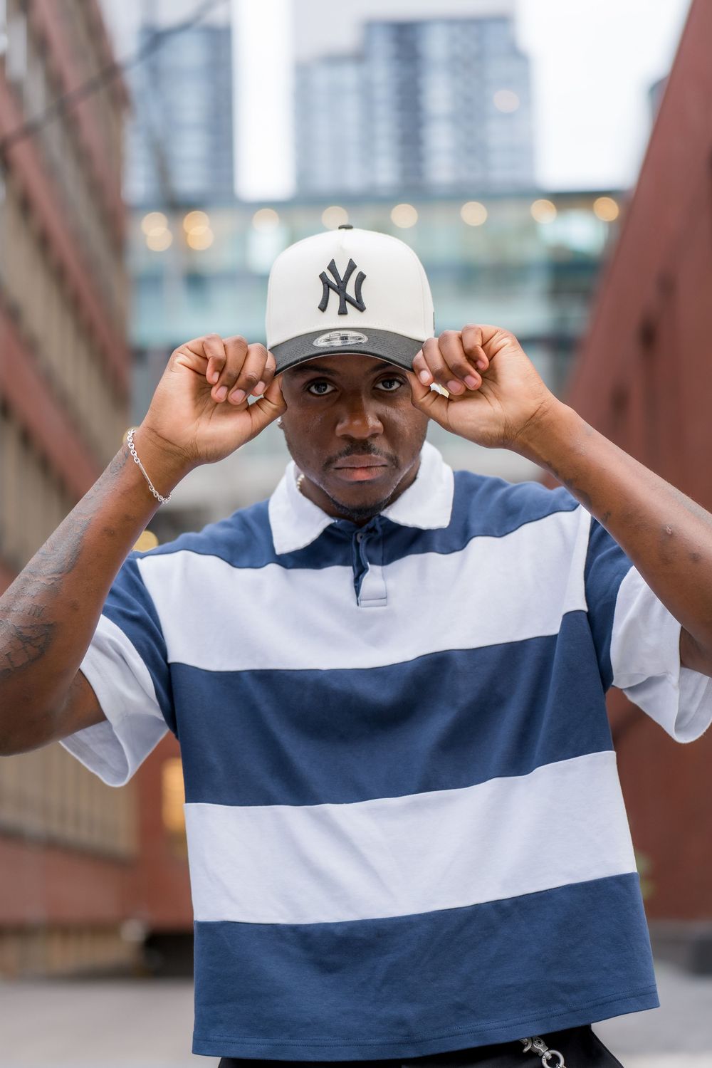 Young Black man adjusting NY Yankees cap, wearing blue striped polo shirt, posing outdoors in urban setting.