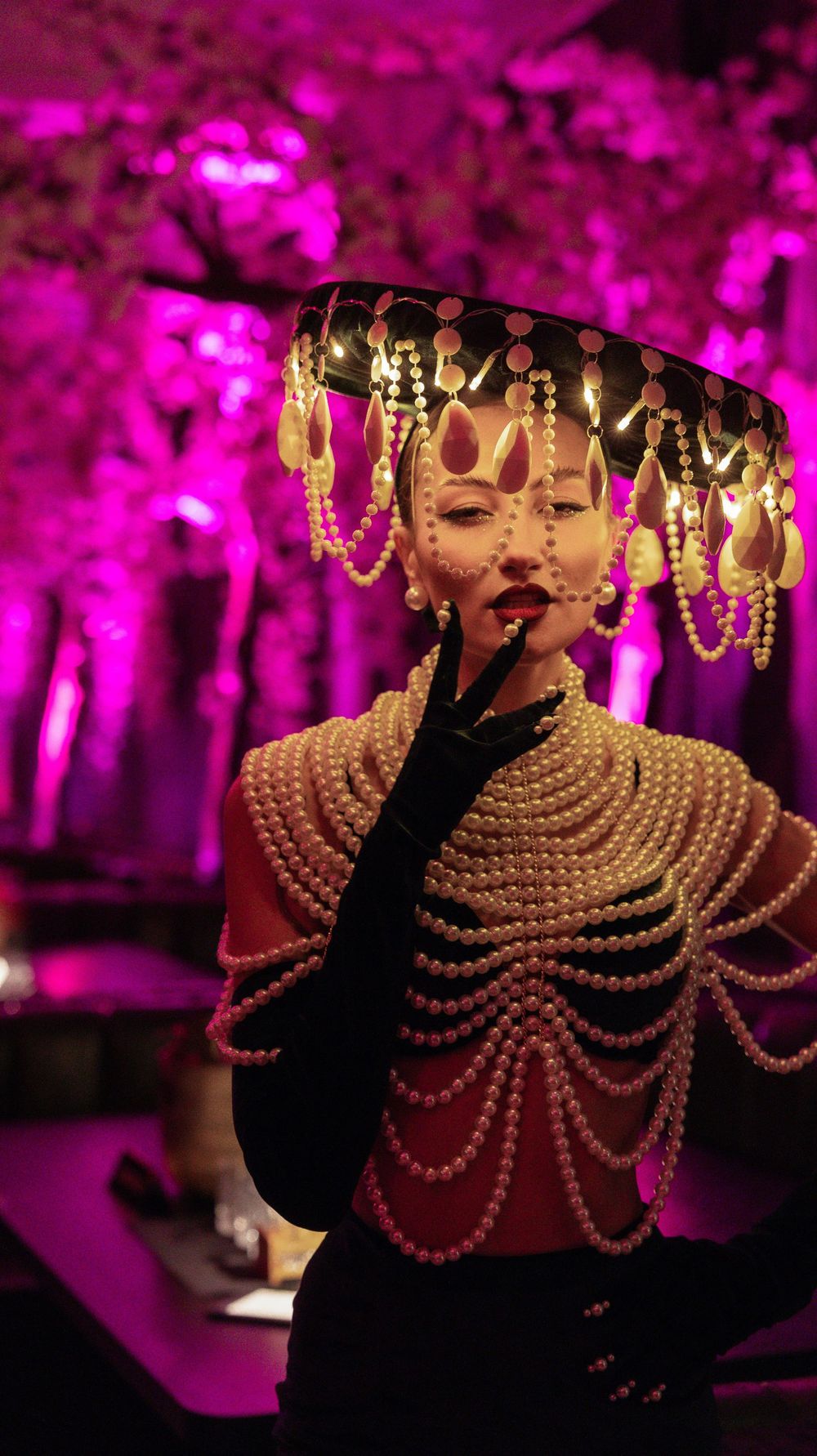 Woman in elaborate pearl costume and ornate headdress poses against vibrant pink lit backdrop.