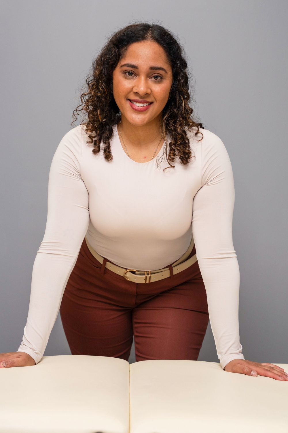 Smiling woman in white long-sleeve top and burgundy pants leaning on a massage table in a studio.