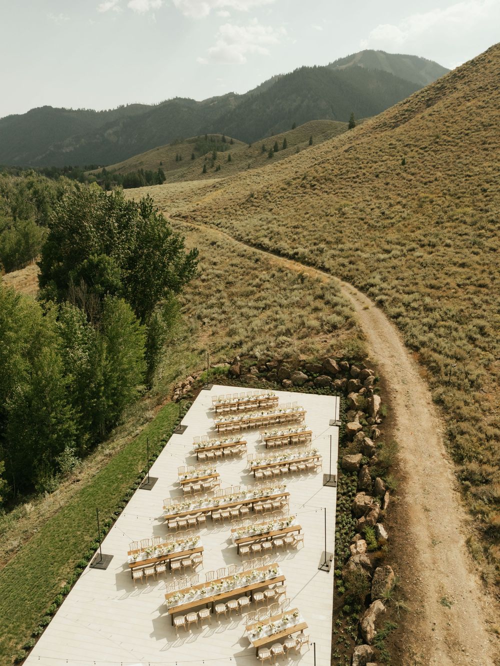 Outdoor wedding ceremony setup with rows of wooden chairs on white aisle runner along mountain dirt trail with scenic views.