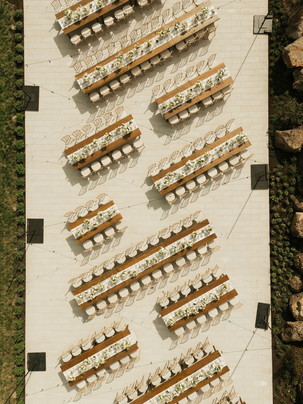Aerial view of wooden banquet tables arranged in diagonal rows on a light-colored stone patio surrounded by greenery.