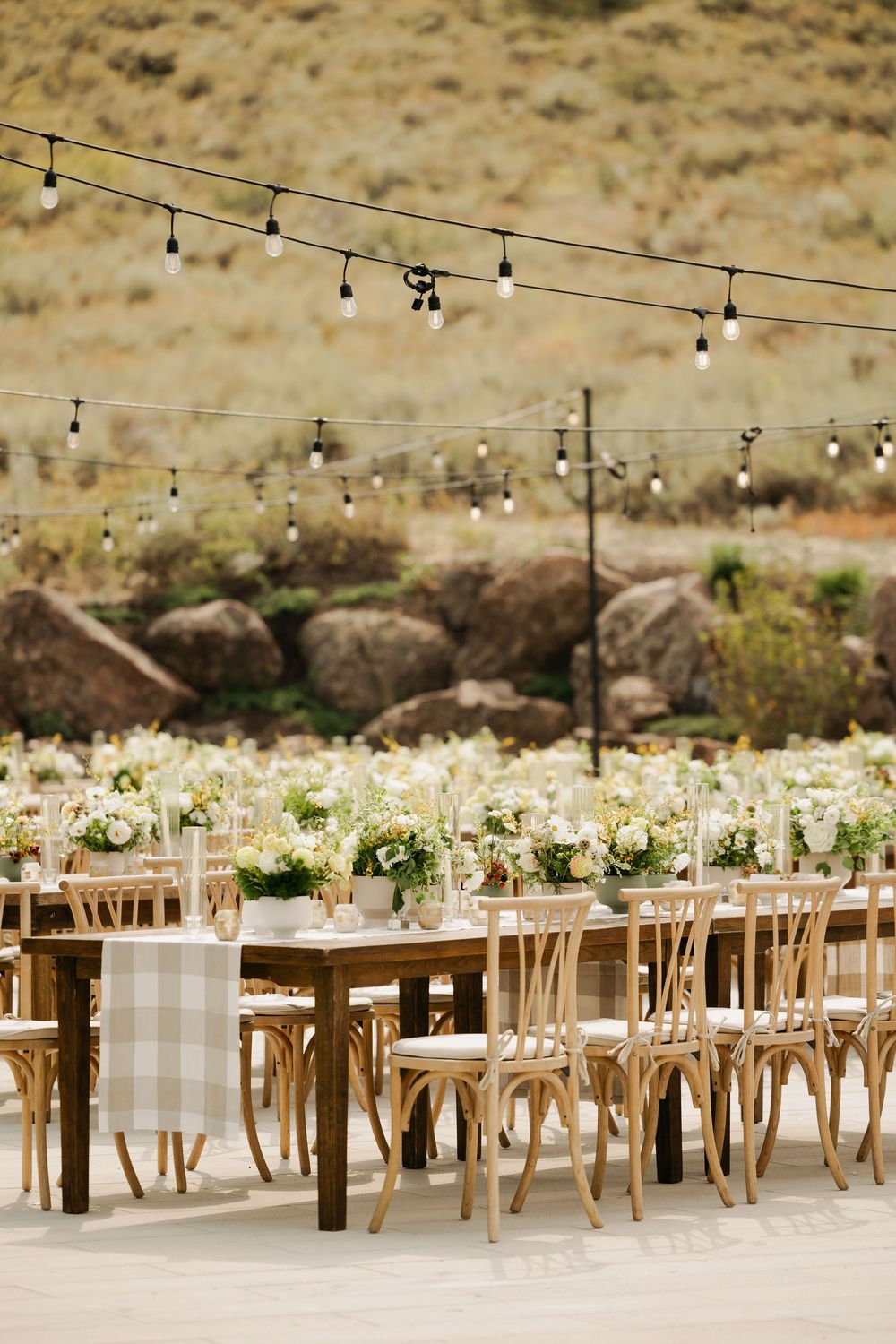 Rustic outdoor wedding reception setup with wooden tables, string lights, and desert mountain backdrop.