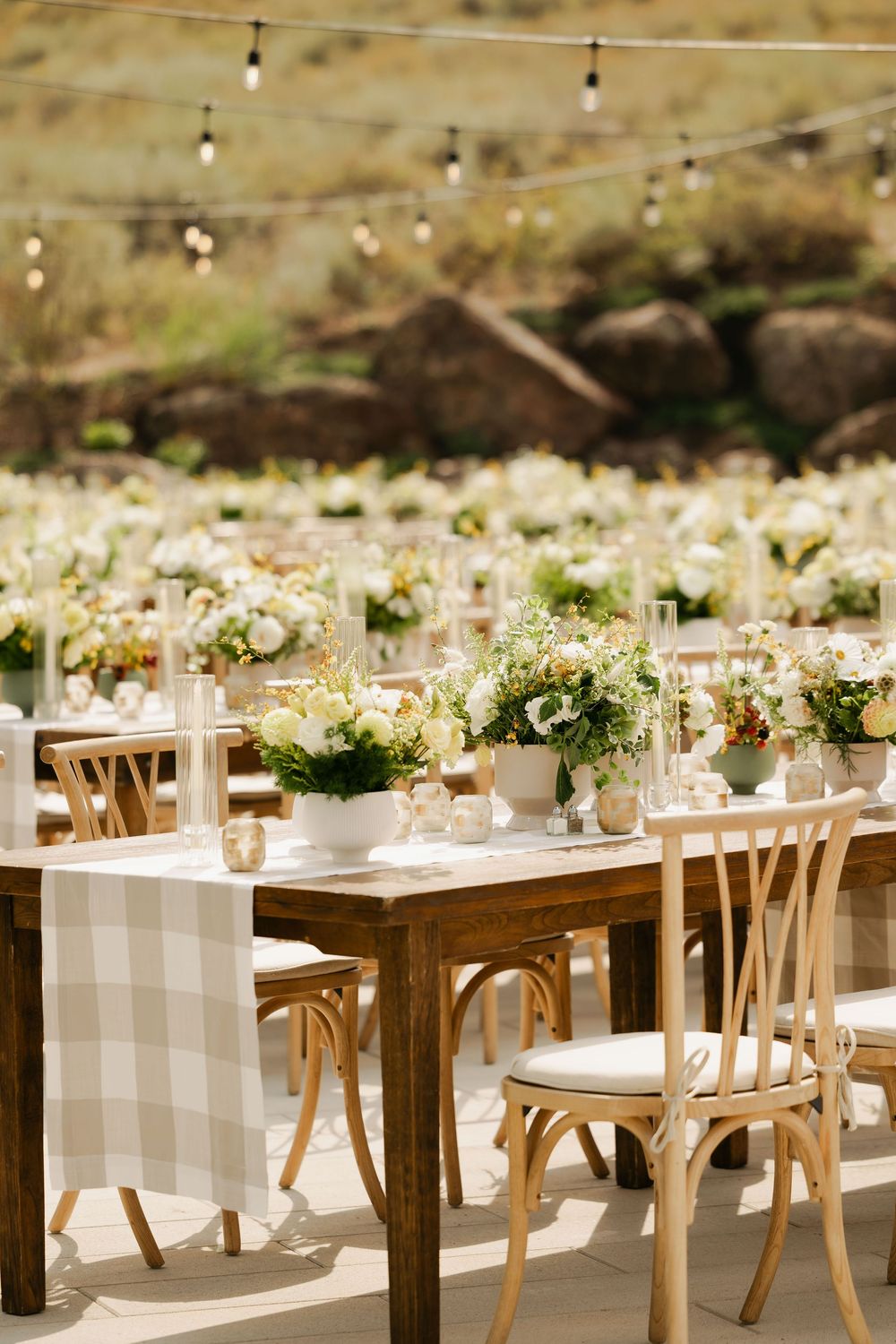 Elegant outdoor wedding reception tables with gingham tablecloths, wooden chairs and white floral centerpieces.