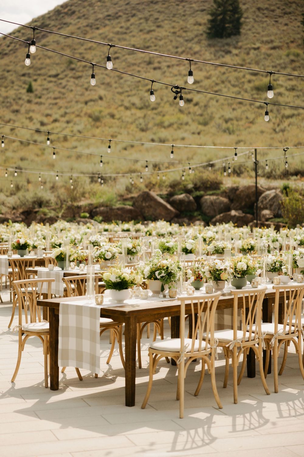 Elegant outdoor wedding reception setup with wooden tables and string lights against a desert mountain backdrop.