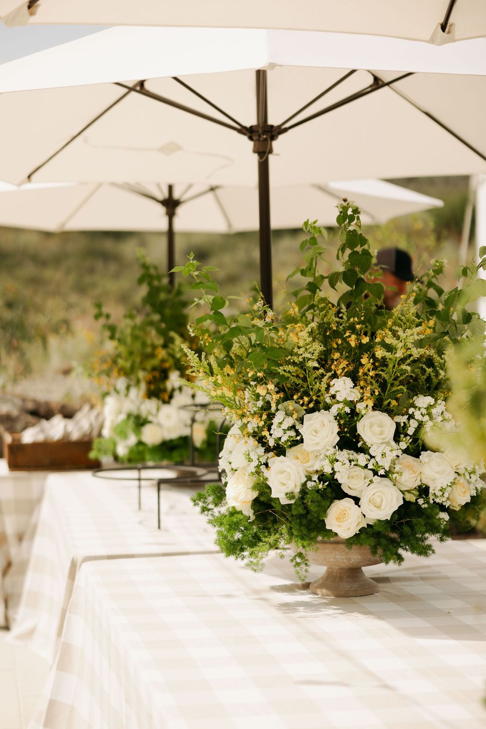 Elegant white floral centerpieces with greenery arranged under white umbrellas at an outdoor event.