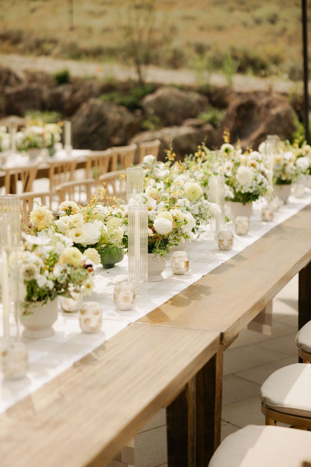 Elegant wedding table setting with white floral centerpieces, candles and rustic wooden tables at an outdoor venue.
