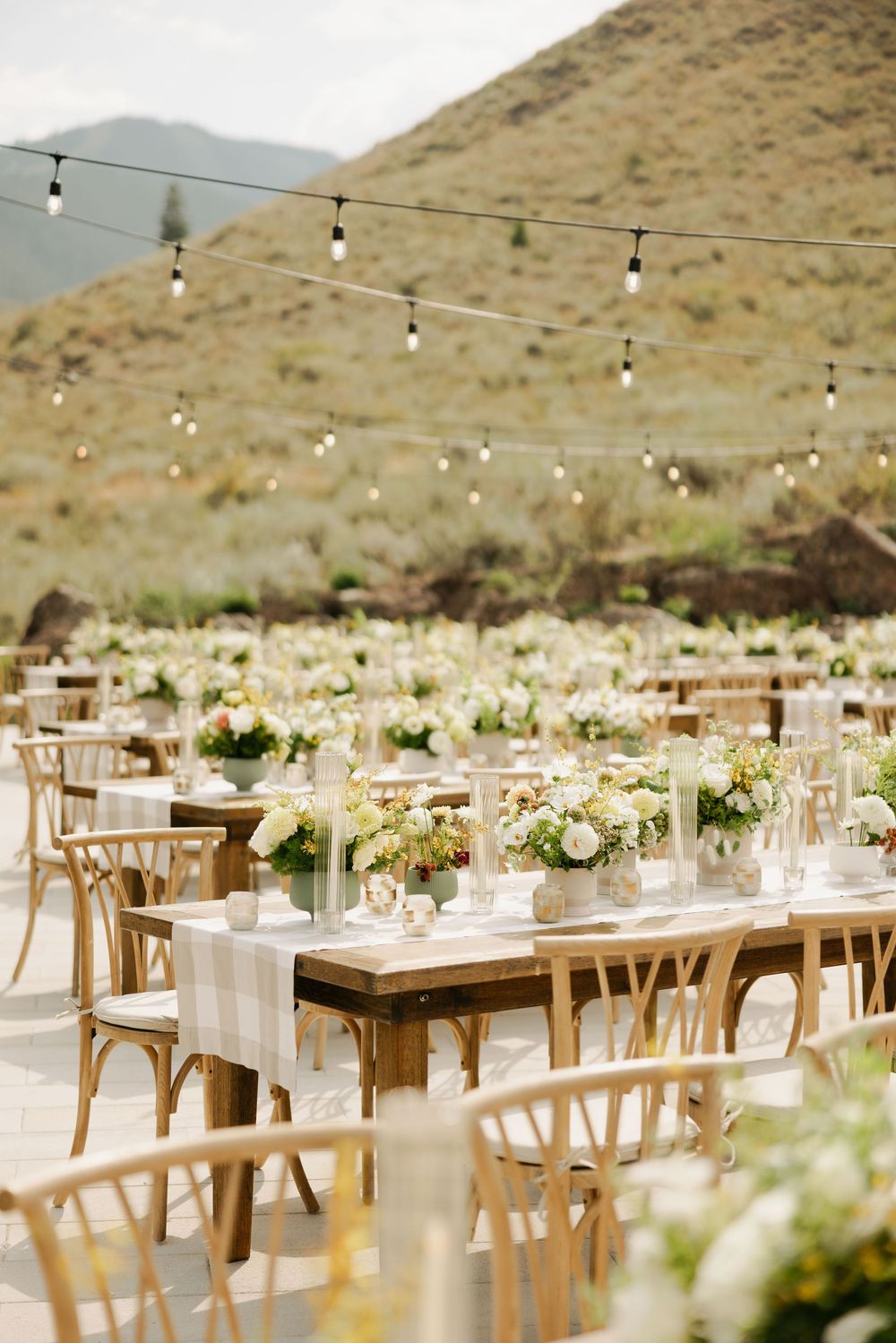 Elegant outdoor wedding reception setup with wooden tables, string lights and mountain views in a desert landscape.